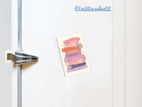 A white fridge with a chrome handle showcases Onderkast Studio’s Smut Book Stack Refrigerator Magnet—a pastel stack of playful, smutty book titles—perfect for naughty bookworms, with Elektrofit on a blue magnet above.