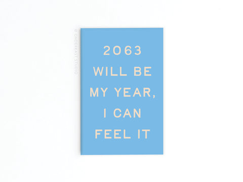 A blue, vibrantly printed refrigerator magnet from Onderkast Studio with beige text reading 2063 will be my year, I can feel it is displayed against a plain white background—perfect for a touch of futuristic outlook.
