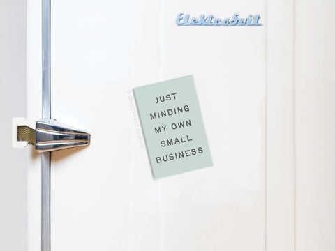 A Just Minding My Own Small Business refrigerator magnet from Onderkast Studio is attached to a white fridge, making it a fun and inspiring accent for entrepreneurs.