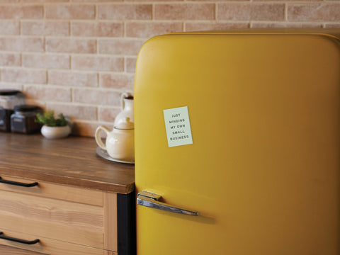 A yellow retro-style fridge features the Onderkast Studio Just Minding My Own Small Business refrigerator magnet, ideal for entrepreneurs. It’s set in a kitchen with a wooden countertop, jars, teapot, and brick backsplash.