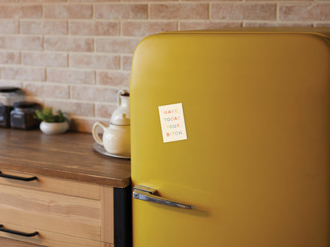 A cozy kitchen with wooden countertops, jars, and a teapot against a brick wall features a yellow retro fridge adorned with the Make Today Your Bitch Refrigerator Magnet by Onderkast Studio.