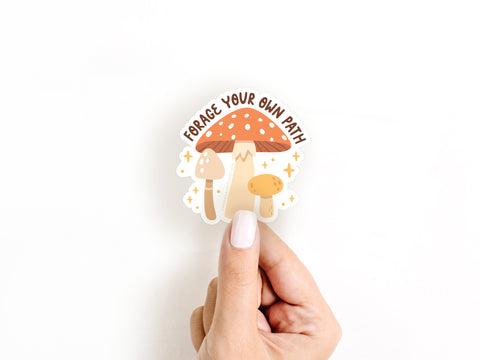 A hand with white nail polish holds Onderkast Studio’s Forage Your Own Path Mushroom Sticker, which features three cartoon mushrooms, brown text, and yellow stars.