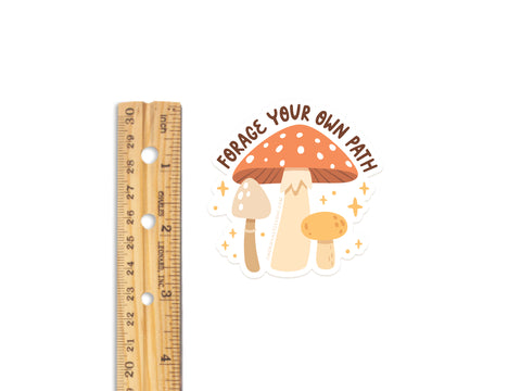 The Forage Your Own Path Mushroom Sticker by Onderkast Studio features three illustrated mushrooms and the phrase FORAGE YOUR OWN PATH. This durable vinyl sticker has a white background and is shown next to a wooden ruler for scale.