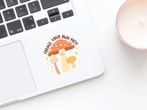 A laptop features the Onderkast Studio Forage Your Own Path Mushroom Sticker, vibrantly printed with three cartoon mushrooms and the phrase FORAGE YOUR OWN PATH, next to a white candle.
