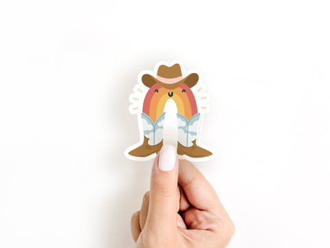A hand with long, white-painted nails holds Onderkast Studio’s Rainbow Cowboy Sticker, featuring a smiling rainbow in a cowboy hat and boots above blue clouds on a white background.