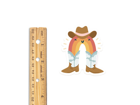 A wooden ruler sits beside the Rainbow Cowboy Sticker by Onderkast Studio; this water-resistant vinyl sticker, about 3 inches tall, features a smiling rainbow with a cowboy hat, boots, jeans, and clouds on a white background.