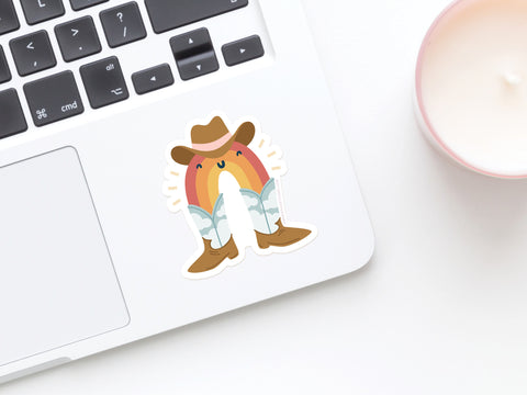 A laptop with the Rainbow Cowboy Sticker by Onderkast Studio—a smiling rainbow with a cowboy hat and boots—on the keyboard sits next to a white candle in a pink holder.
