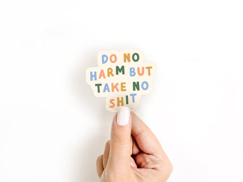 A hand with white-painted nails holds Onderkast Studio’s Do No Harm But Take No Shit Sticker, featuring bold, colorful lettering on weatherproof vinyl against a white background.