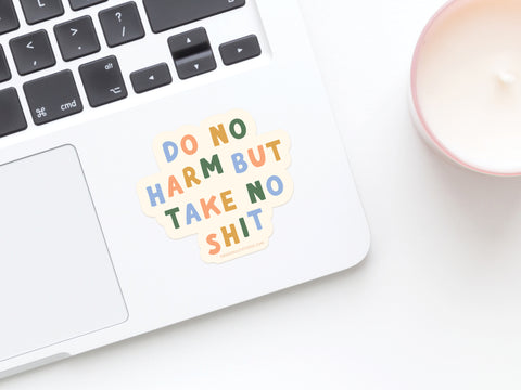 A laptop featuring the Onderkast Studio Do No Harm But Take No Shit Sticker, crafted from weatherproof vinyl, sits next to a lit candle in a pink holder.