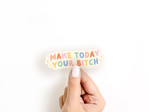 A hand with white nails holds the colorful Make Today Your Bitch Sticker by Onderkast Studio, featuring bold letters on a white background. This vibrant sticker is proudly printed in the USA.