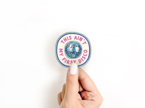 A hand with white nail polish holds the Onderkast Studio This Ain’t My First Disco Glitter Sticker, featuring a disco ball and pink and purple text on weatherproof white vinyl.