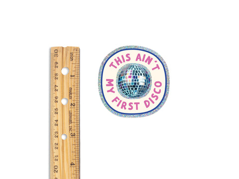 A wooden ruler beside the Onderkast Studio “This Ain’t My First Disco Glitter Sticker,” a weatherproof vinyl sticker featuring a shiny disco ball and text, measuring about 2 inches wide.