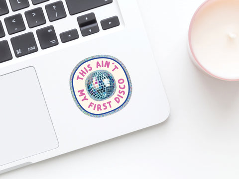 A silver laptop keyboard is decorated with Onderkast Studio’s “This Ain’t My First Disco Glitter Sticker”—a weatherproof vinyl, round sticker featuring a disco ball and the phrase—while a white candle in a pink holder sits nearby.