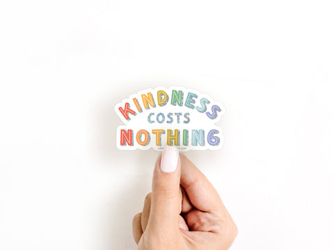 A hand with white nail polish holds an Onderkast Studio Kindness Costs Nothing Glitter Sticker made from weatherproof vinyl, shown against a white background.