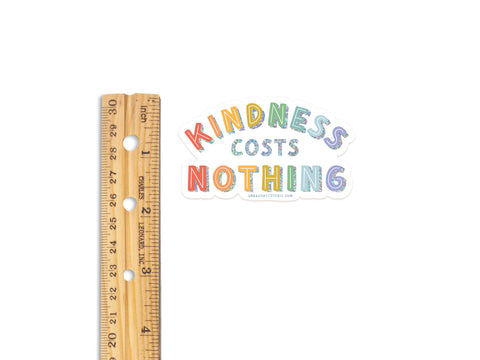 A wooden ruler is next to an Onderkast Studio Kindness Costs Nothing Glitter Sticker, each word in a playful font, on a white background. This weatherproof vinyl sticker adds cheerful inspiration wherever you place it.