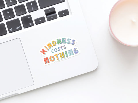 A laptop with a vibrant Onderkast Studio Kindness Costs Nothing Glitter Sticker, made from weatherproof vinyl, sits by a white candle in a light pink holder on a white surface.