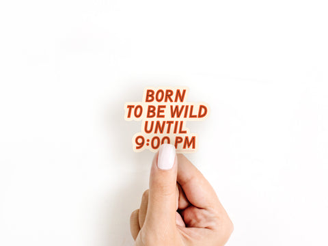 A hand with white-painted nails holds the Born To Be Wild Sticker by Onderkast Studio, a bold weatherproof vinyl sticker with red and orange lettering on a crisp white background.