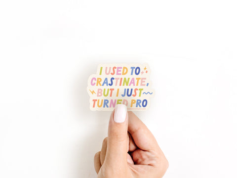 A hand with light skin and white nail polish holds the Onderkast Studio Crastinator Pro Sticker, which features “I used to procrastinate, but I just turned pro.” Made from weatherproof vinyl. Shown against a plain white background.