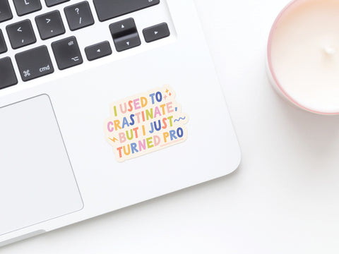 A laptop with the Crastinator Pro Sticker by Onderkast Studio in weatherproof vinyl reads, “I used to procrastinate, but I just turned pro.” A pink candle sits nearby on the white surface.
