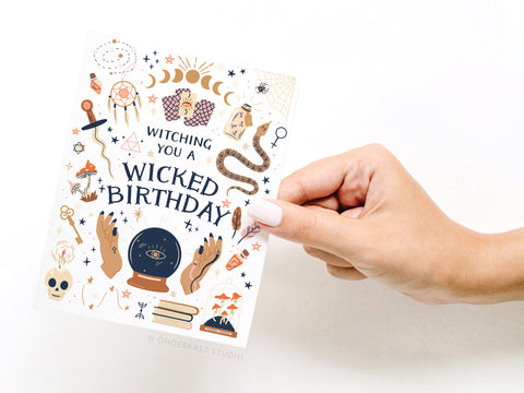 A hand with light pink nails holds the Witching You a Wicked Birthday Greeting Card by Onderkast Studio, featuring magical witch-themed illustrations and bold, enchanting lettering.