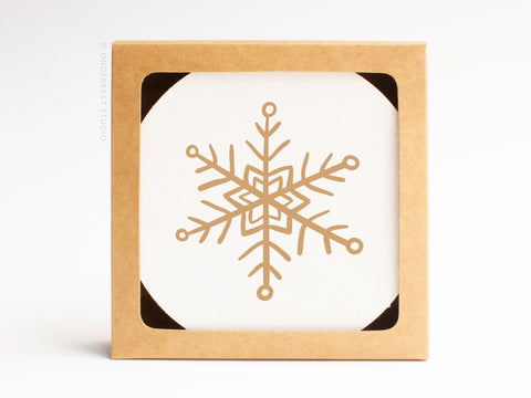 The Snowflake Coaster Set by Onderkast Studio is presented in a square box with a cutout, showcasing a white coaster adorned with a gold geometric snowflake for an elegant, handmade touch.