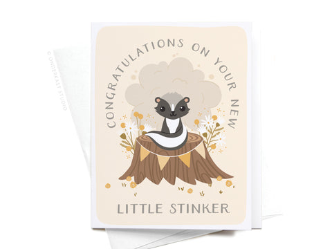 Onderkast Studios Congratulations On Your New Little Stinker Greeting Card features a cute cartoon skunk on a tree stump with flowers and the message: “Congratulations on your new little stinker” on a light beige background.