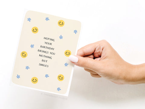 A hand holds the Nothing But Smiles Greeting Card by Onderkast Studio, decorated with yellow smiley faces and blue flowers. The card reads: Hoping your birthday brings you nothing but smiles.