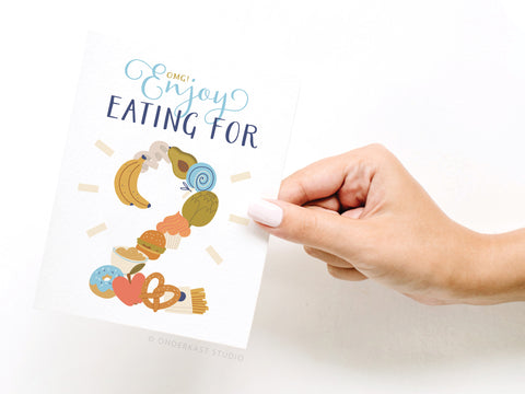 A hand with light-pink nails holds the Onderkast Studio OMG! Enjoy Eating for 2 Greeting Card—FINAL STOCK. Whimsical foods form the number two, making it perfect for expecting parents!.