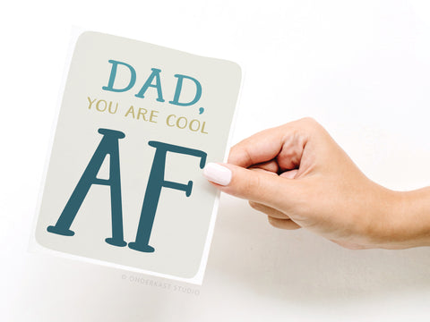 A hand with white-painted nails holds the Onderkast Studio Dad, You Are Cool AF Greeting Card featuring bold, colorful text—perfect for celebrating dad’s cool vibes this Fathers Day.