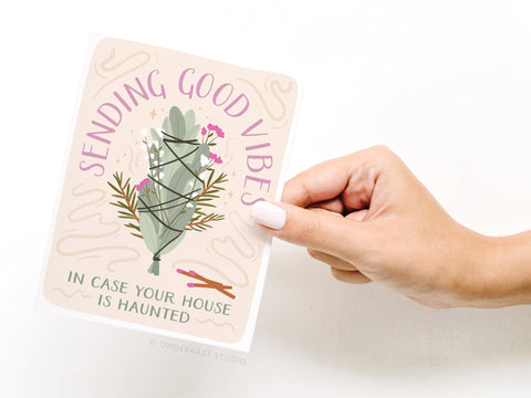 A hand with light pink nail polish holds the Onderkast Studio Sending Good Vibes Smudge Stick Greeting Card, which reads, Sending good vibes in case your house is haunted, with a sage and flower illustration for positivity.