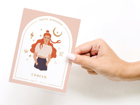 A hand holds the Happy Birthday Cancer Zodiac Greeting Card by Onderkast Studio, featuring an illustrated woman with long red hair, zodiac symbols, and the text “Happy Birthday Cancer, June 21 - July 22” on a pale pink background.