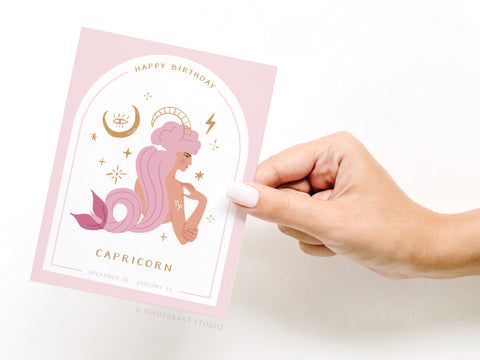 A hand with pale pink polish holds the Happy Birthday Capricorn Zodiac Greeting Card by Onderkast Studio, featuring an illustrated Capricorn with purple hair amid stars and zodiac symbols, plus “Happy Birthday Capricorn.”.