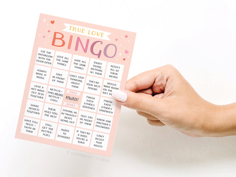 A hand holds the True Love Bingo Greeting Card by Onderkast Studio, featuring hearts, playful typography, and fun couple activities on a pink background—perfect for gifting to your special someone.