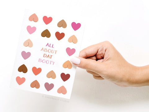 A well-groomed hand displays the All About Dat Booty Greeting Card by Onderkast Studio, featuring heart shapes in diverse skin tones and the phrase ALL ABOUT DAT BOOTY at the center.