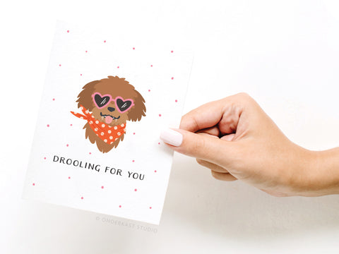 A manicured hand holds the Onderkast Studio Drooling For You Dog Greeting Card, which features a cartoon pup in heart-shaped sunglasses and an orange polka dot bandana, with DROOLING FOR YOU printed below.