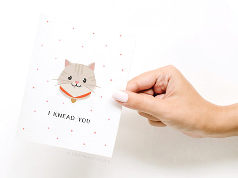 A hand with light pink nail polish holds the Onderkast Studio “I Knead You Cat Greeting Card,” featuring a cute cartoon cat with a collar, vibrant colors, small pink dots, and the phrase I KNEAD YOU to share feline affection.