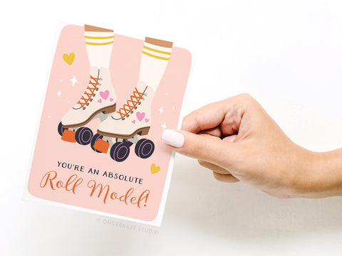 A hand holds Onderkast Studio’s Roll Model Roller Skates Greeting Card, featuring heart-accented skates, yellow-striped socks, and the message “You’re an absolute Roll Model!” with a nostalgic background of tiny hearts.