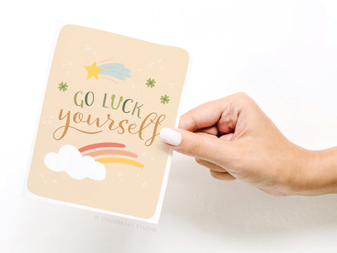 A hand with light pink nails holds the Go Luck Yourself Greeting Card – FINAL STOCK by Onderkast Studio, featuring “Go Luck Yourself,” stars, a shooting star, rainbow, and cloud on a premium beige background.