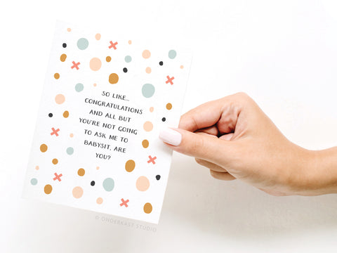 A manicured hand holds Onderkast Studio’s “Don’t Ask Me to Babysit” Greeting Card, adorned with pastel dots and crosses. The front reads: So like... congratulations and all but you’re not going to ask me to babysit, are you?.