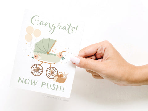 A hand holds Onderkast Studio’s Now Push! Baby Stroller Greeting Card, featuring a vintage stroller, balloons, basket, and flowers with Congrats! NOW PUSH! in green and beige—a charming gift for new parents.