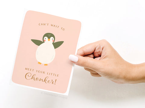 A hand with pale pink nail polish holds Onderkast Studio’s “Can’t Wait to Meet Your Little Chonker” Greeting Card – FINAL STOCK, featuring a cute illustrated penguin on a pink background.