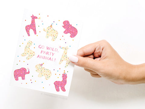 A hand holds the Onderkast Studio Go Wild Party Animal Frosted Cookies Greeting Card, adorned with colorful, polka-dotted animals in party hats and the message “GO WILD, PARTY ANIMAL!”—a perfect choice for birthday celebrations.