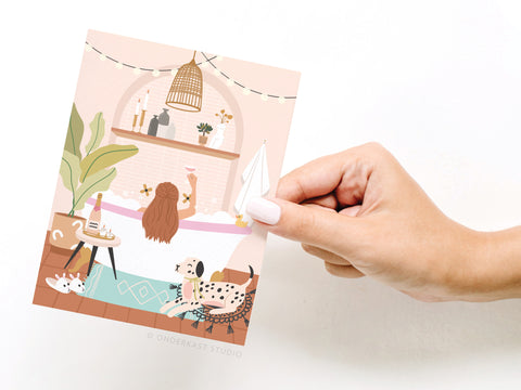 A hand holds the Bubble Bath Cheers Greeting Card by Onderkast Studio, featuring an illustrated woman relaxing in a bubble bath with wine, plants, candles, pets, and cozy decor in a stylishly lit bathroom.