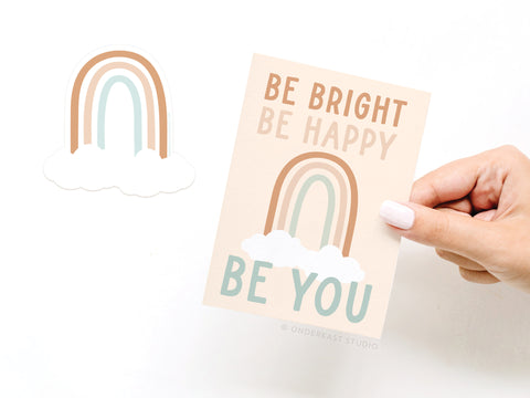 A hand holds Onderkast Studio’s Be Bright Be Happy Be You Rainbow Sticker Greeting Card, featuring a pastel rainbow, clouds, and uplifting words. A matching vinyl sticker rests beside the card on a white background.