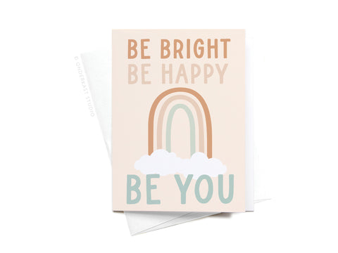 The Be Bright Be Happy Be You Rainbow Sticker Greeting Card by Onderkast Studio features a pastel rainbow and clouds with the uplifting phrase in light brown and teal, displayed atop a white envelope.