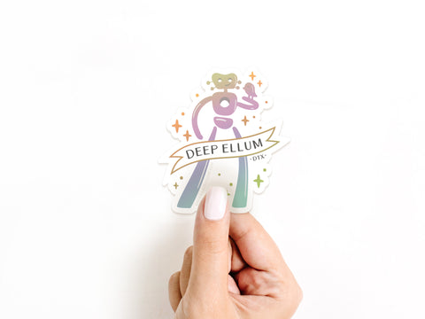A hand with light skin and white nail polish holds the Deep Ellum Traveling Man Holographic Sticker by Onderkast Studio. The durable vinyl shines with colorful abstract art, stars, and a “DEEP ELLUM - DTX-” banner on a white background.