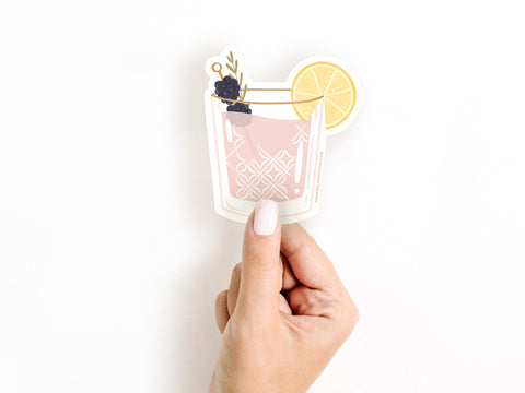 A hand with pale nail polish holds the Onderkast Studio Berry Cocktail Sticker—a durable, water-resistant vinyl sticker featuring a pink drink with a lemon slice and two blackberries—against a plain white background.