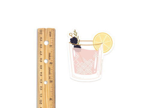 A wooden ruler beside the Onderkast Studio Berry Cocktail Sticker shows the water-resistant vinyl sticker—featuring a pink drink with lemon and blackberries—is about 3 inches tall.