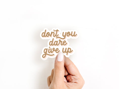 A hand with light nail polish holds the Onderkast Studio Dont You Dare Give Up sticker, a durable, water-resistant vinyl decal featuring brown cursive text on a white background.