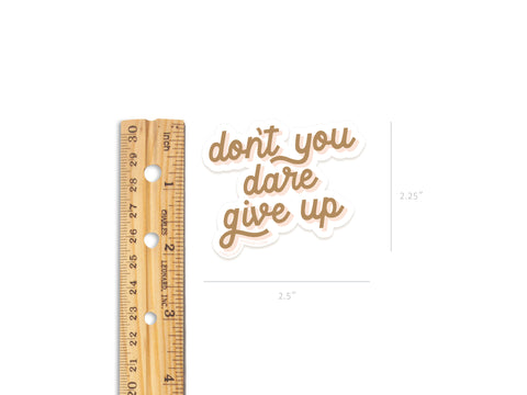 A wooden ruler lies next to Onderkast Studio’s “Don’t You Dare Give Up Sticker,” a durable, water-resistant vinyl sticker in brown script measuring 2.5 x 2.25, shown on a white background.